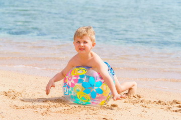 Smiley little blond kid is playing with the inflatable translucent beach ball on the sandy sea or ocean shore. Hard sun light summer day.