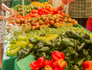Farmer's market produce for sale at stand. Color, horizontal photo of heirloom tomatoes, peppers, brussel sprouts, squash. Woman's hand on left holding a tomato, man reaching for tomatoes on right.