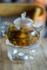 Glass kettle with blooming flower tea on wooden table. Stand with little candle. Bundle of flowering green tea inside teapot. Chinese tea making tradition. Blurred background. For cafe, restaurant.