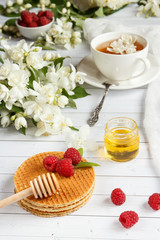 Tea Cup with Jasmine flowers and thin waffles with honey and raspberry on a light wooden background