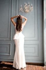 Невеста с красивой фигурой стоит спиной в белом длинном платье в интерьерной студии 