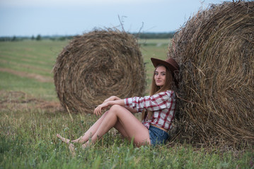 Naklejka na ściany i meble Young woman in shorts and cowboy hat sitting near a haymow