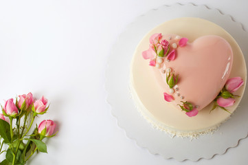 Obraz na płótnie Canvas Two-tiered modern french mousse cake with pink mirror glaze in the shape of a heart decorated with roses on a white background. Picture for a menu or a confectionery catalog. Top view. Text space.