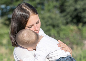 Young beautiful mother and baby boy hugging on nature in summer. Pretty woman Holding sleeping child ion her arms