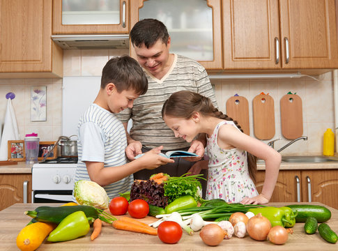 Father and two children reading cooking book and choice dishes. Happy family, girl and boy having fun with fruits and vegetables in home kitchen interior. Healthy food concept.