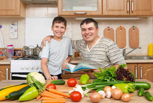 Father and child reading cooking book and choice dishes. Happy family having fun with fruits and vegetables in home kitchen interior. Healthy food concept.