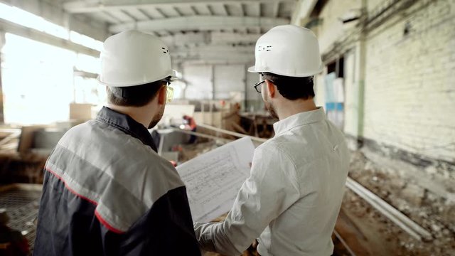 Back view of men in helmets and eyeglasses standing together in construction area with people in the background. Architect and worker looking at structure plan and discussing details of development.