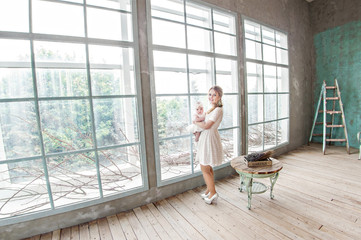 A beautiful mother in a white dress is standing by the big window and is holding a little girl in her arms and smiling