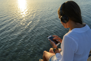 Girl is sitting on a pier by the sea in the sun lights with smartphone in headphones and listening to music