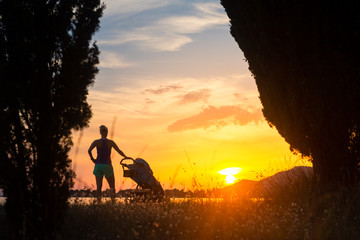 Fototapeta premium Silhouette of mother with stroller enjoying motherhood at sunset landscape