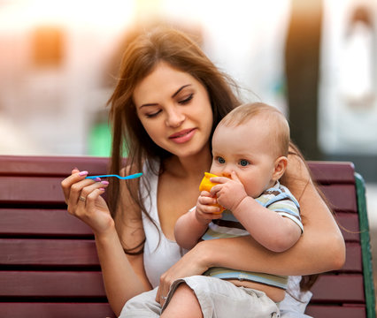 Baby feeding spoon by mother in park outdoor. Weaning in first weeks. Eating child summer on bench. Good appetite by baby food with foster mother number one. Color tone on shiny sunlight background.