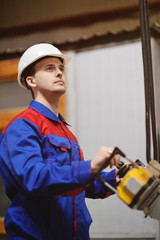 A young handsome worker in a white helmet with a large remote control in the background of the plant.