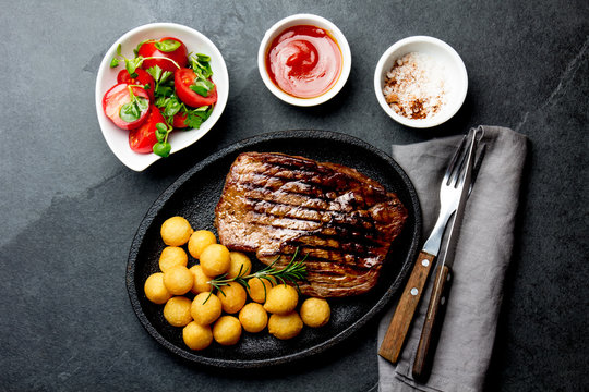 Grilled beef steak served on cast iron plate with tomato salad and potatoes balls. Barbecue, bbq meat beef tenderloin. Top view, slate background