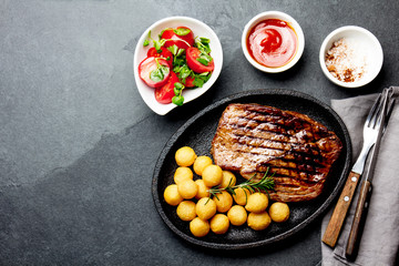 Grilled beef steak served on cast iron plate with tomato salad and potatoes balls. Barbecue, bbq meat beef tenderloin. Top view, slate background