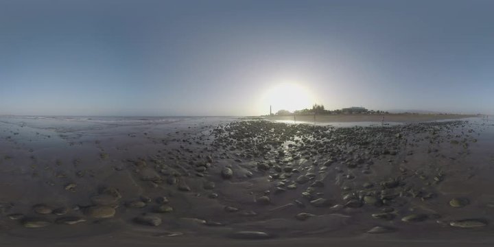 GRAN CANARIA, CANARY ISLANDS - AUGUST 02, 2016: 360 VR Video. People on vacation walking along the ocean coast with rocks. View to the resort with Maspalomas Lighthouse against setting sun