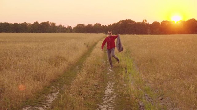 Successful businessman runs skipping along a country road near a wheat field at sunset, waving his jacket in slow motion