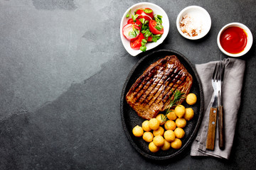 Grilled beef steak served on cast iron plate with tomato salad and potatoes balls. Barbecue, bbq meat beef tenderloin. Top view, slate background