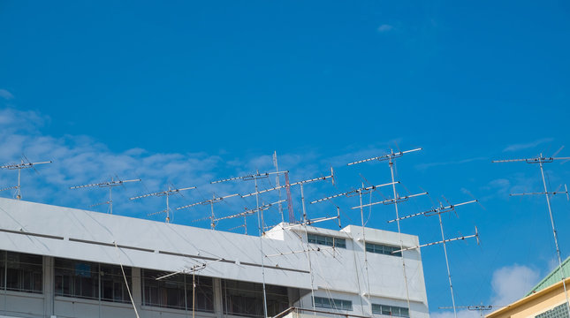 Many television antennas on the roof of building with blue sky.