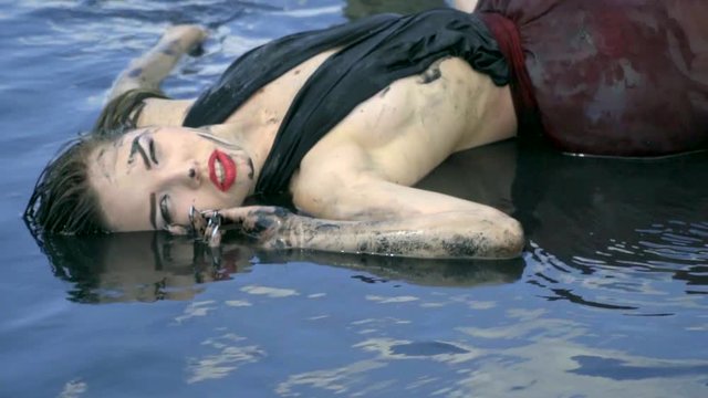 A beautiful blonde smeared in mud and a wet dress lies in the estuary, licking water from her lips and resting