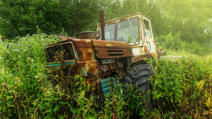 An old rusty derelict tractor parked in a farm yard in amongst overgrown grass and weeds