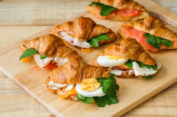 Croissant sandwiches served on wooden board. Croissants with egg, melted cheese, parsley, chicken ham, smoked salmon