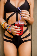 Portrait of a sexy crazy thirsty slim lady in red wig, black sensual swimsuit and fashion sunglasses with glass of red soda water. Beauty cute girl on a tropical resort. Outdoor summer lifestyle. - 164802522