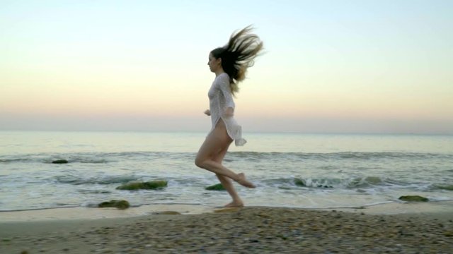 Young girl in short white dress runs along the beach of the sea against the sunset background. Slow motion shooting