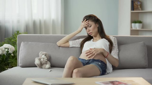 Woman pregnant with child touching belly and head, looking preoccupied, worries