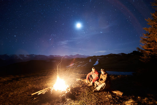 Two male tourists have a rest in his camp near the forest at night. Guys sitting near campfire and tent under beautiful night sky full of stars and the moon, and enjoying night scene in the mountains