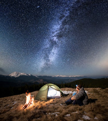 Male tourist have a rest in his camp at night. Man with a headlamp sitting near campfire and tent under beautiful sky full of stars and milky way. On the background snow-covered mountains and forests © anatoliy_gleb