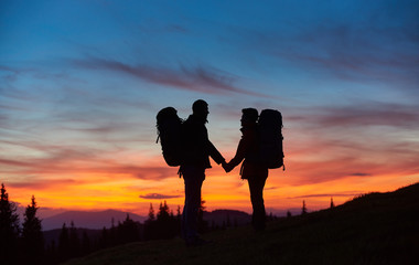 Happy couple hiking together holding hands standing with their backpacks on top of a mountain during stunning sunset people love silhouettes meadow active relationships