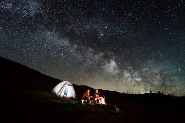 Night camping. Romantic couple tourists enjoying at a campfire near illuminated tent under beautiful night sky full of stars and milky way. Long exposure