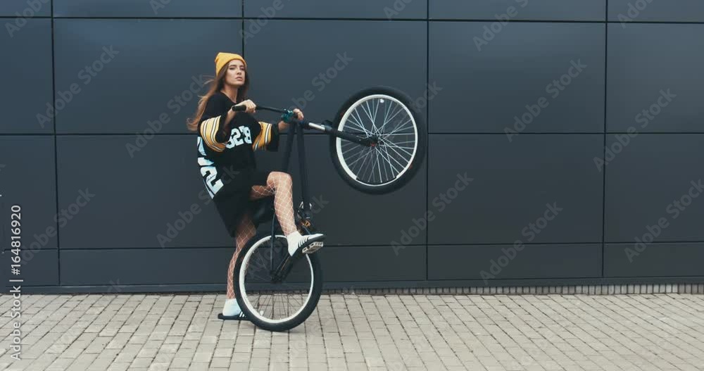 Wall mural Young stylish Caucasian female sitting on a bicycle against grey wall background. 4K UHD RAW edited footage