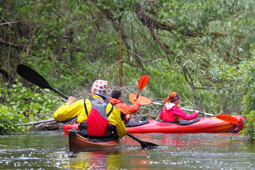 Man in selfmade brown wooden kayak in red and yellow life jacket and friends kayaking in wild river among thickets of plants on biosphere reserve in spring