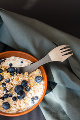 Healthy breakfast. Oat granola with fresh blueberries, currants and milk in a clay bowl over dark grunge surface. Top view, copy space. Metal spoon with berries