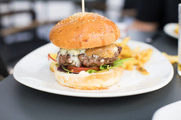still life of white dish with beef burger, with cheese, onion, pickle, lettuce and tomato slice, inside bun bread and potatoes french fries on dark table at cafe restaurant
