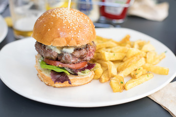 still life of beef burger, with cheese, onion, pickle, lettuce and tomato slice, inside bun bread and potatoes french fries on dark table at cafe restaurant
