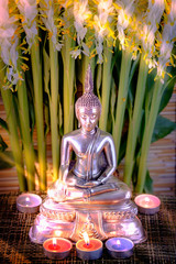 Selective focus of buddha statue with blurred burning candle light in soft  light and white flower of globba tree. Concept of peace, meditation, hope and relaxation