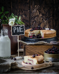 pie with black currant on wooden box with one slice of pie on a plate with metal jar of fresh berries, brown pot with white flowers, bottle of milk and small desk with a word 