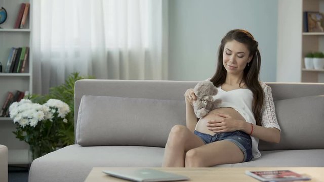Woman pregnant with child putting teddy bear on belly, communication with baby
