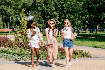Naklejka na ściany i meble happy multicultural women with disposable cups of coffee in hands walking in park