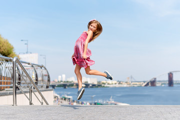 Happy little girl jumping and flying outdoors near the river embankment. Summer vacation and children's games to spend time in the open air
