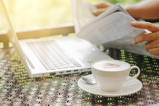 Stock photo :.Cappuccino coffee cup on a vintage table with laptop of a man reading a newspaper in the morning light.