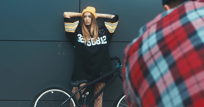 Photographer taking pictures of young stylish Caucasian female posing with a bicycle against grey wall background. 4K UHD RAW edited footage