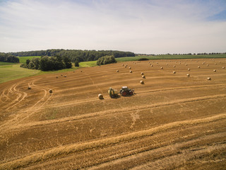 Obraz premium Aerial view of tractor with trailer at working in the golden wheat field - trailer makes straw bales - harvest in the summer - top view - Germany
