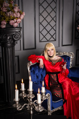 Fototapeta na wymiar Beautiful blond woman sitting on the couch. She is dressed in a luxurious, expensive, red dress.