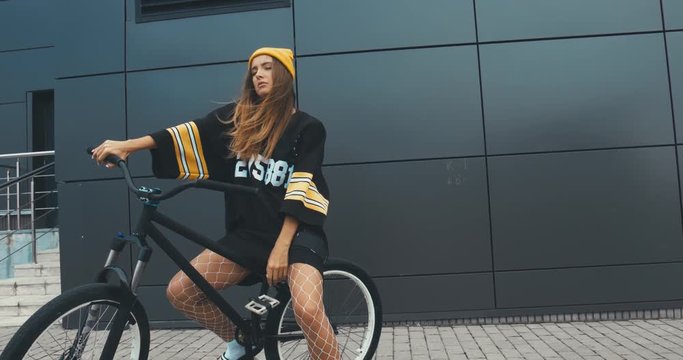 Young stylish Caucasian female in hockey style jersey riding her bicycle against grey wall background. 4K UHD RAW edited footage