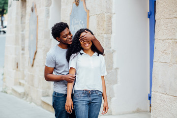 Obraz na płótnie Canvas Beautiful dark skin woman smilling and trying to guess who is standing behind her. Her afro american boyfriend is covering her eyes by hands and smiling. Love and gift concept