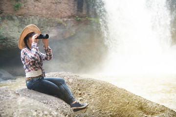 Young Asian traveler woman looking a binoculars in happiness and sitting on a rock of waterfall.