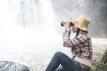 Young Asian traveler woman looking a binoculars in happiness and sitting on a rock of waterfall.